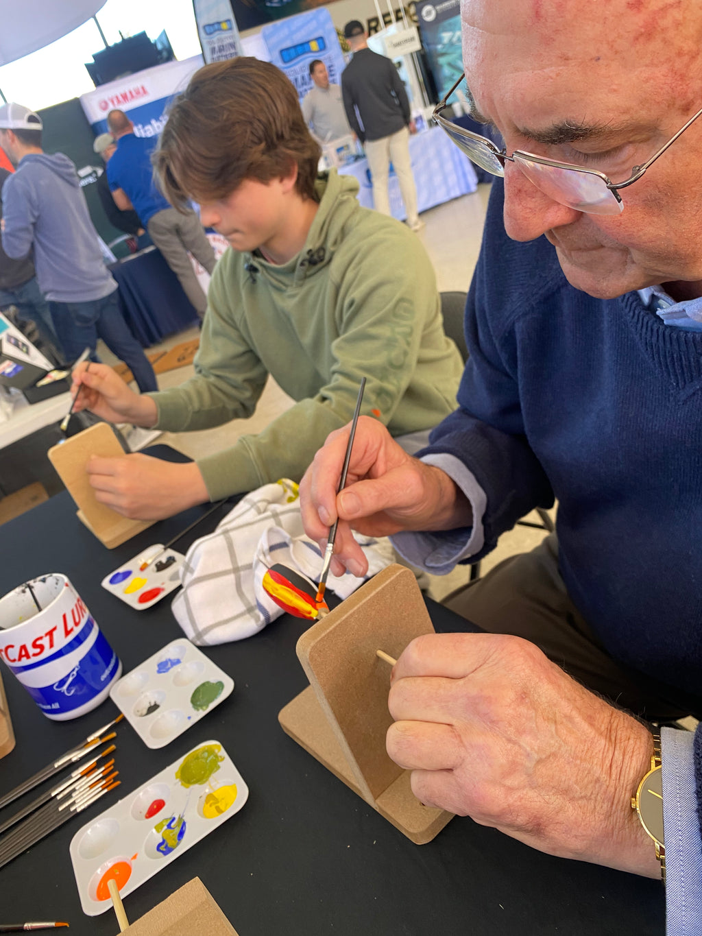 Two people working on a craft project at an event with booths in the background.